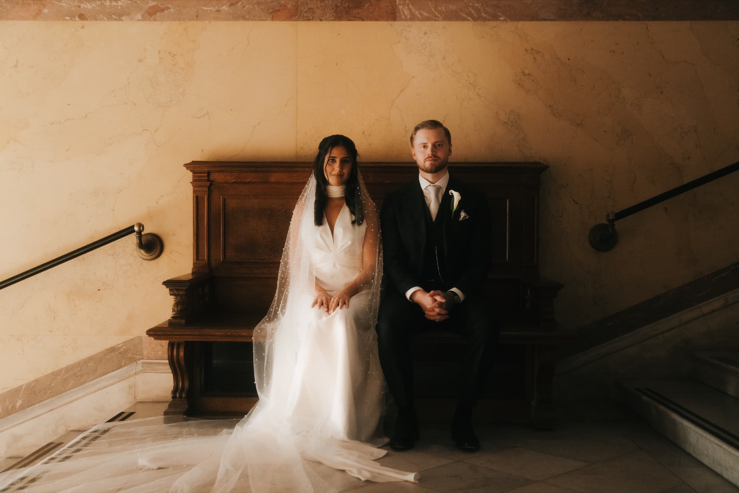 Couple seated together on a wooden bench against a marble wall