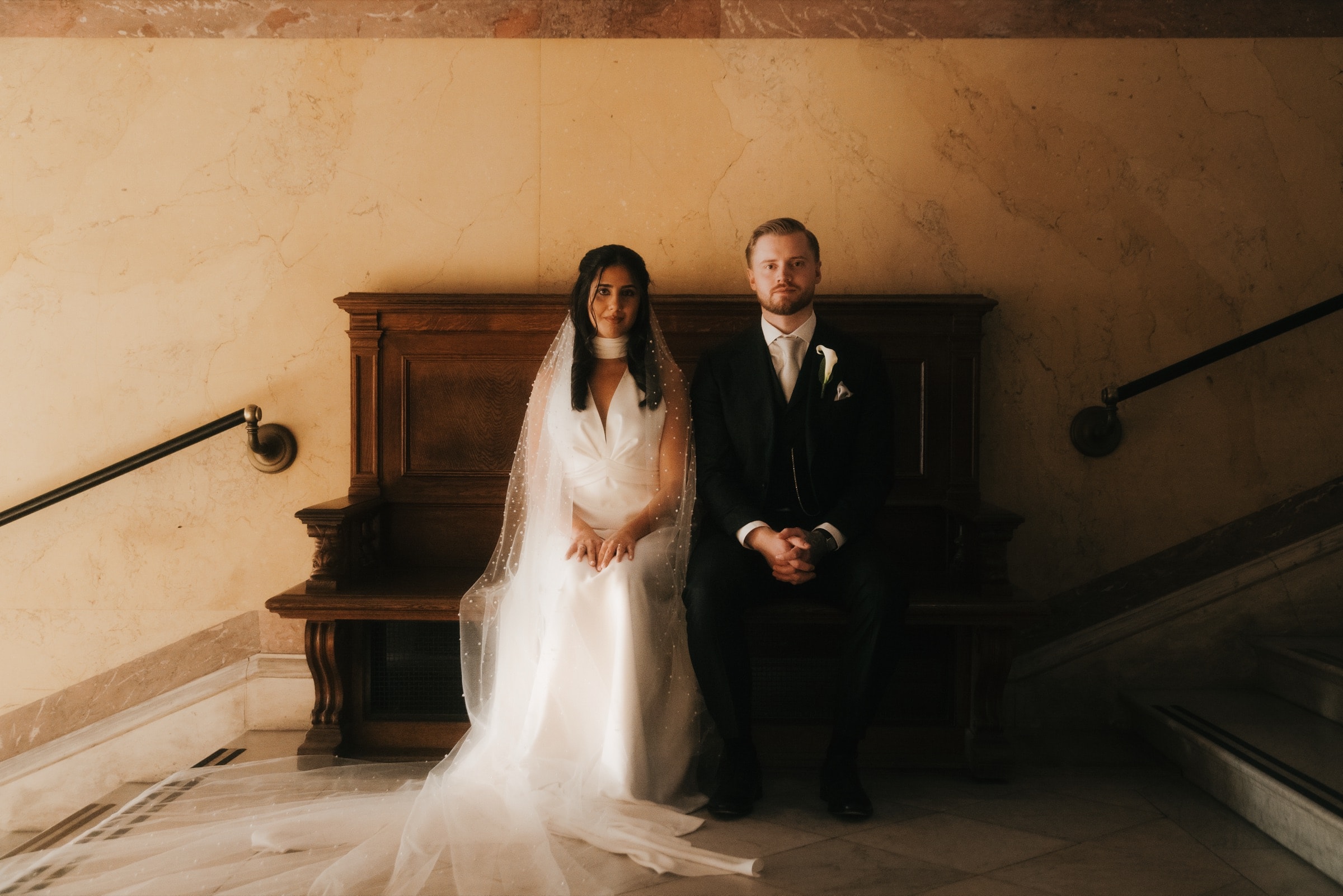 Couple seated together on a wooden bench against a marble wall
